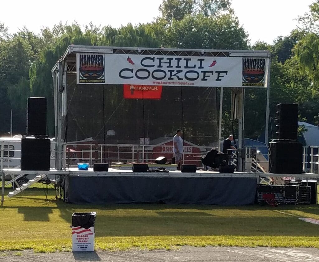 Chili cook-off festival event in New York A view of an outdoor event with chairs and tables. An event stage with an open top and a person walking on the grass to rent in Long Island and Tri state area from Allseasonsstagerentals.com
