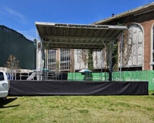 A truck parked in front of an outdoor stage. A view of an outdoor event with chairs and tables. An event stage with an open top and a person walking on the grass to rent in Long Island and Tri state area from Allseasonsstagerentals.com