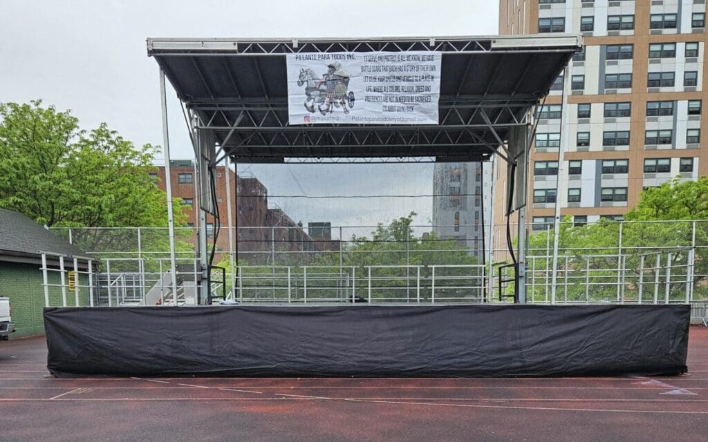 A stage with black tarps on the side of it in New York. A view of an outdoor event with chairs and tables. An event stage with an open top and a person walking on the grass to rent in Long Island and Tri state area from Allseasonsstagerentals.com