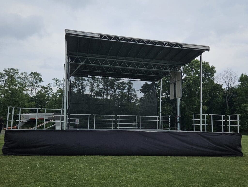 A stage with black tarp covering the top of it.A view of an outdoor event with chairs and tables. An event stage with an open top and a person walking on the grass to rent in Long Island and Tri state area from Allseasonsstagerentals.com