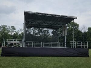 A stage with black tarp covering the top of it.A view of an outdoor event with chairs and tables. An event stage with an open top and a person walking on the grass to rent in Long Island and Tri state area from Allseasonsstagerentals.com