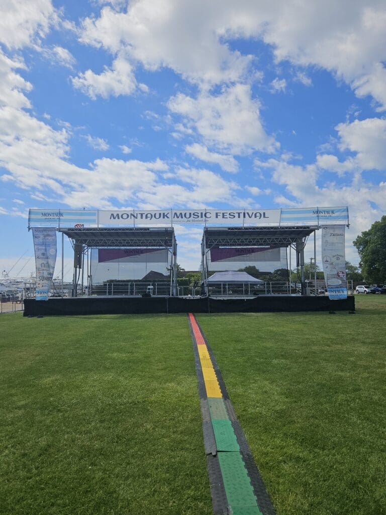 A stage with two levels and a rainbow strip in the middle. A view of an outdoor event with chairs and tables. An event stage with an open top and a person walking on the grass to rent in Long Island and Tri state area from Allseasonsstagerentals.com