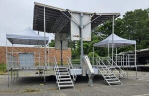 A stage with steps and canopies in the middle of an empty parking lot in Long Island, New York.A view of an outdoor event with chairs and tables. An event stage with an open top and a person walking on the grass to rent in Long Island and Tri state area from Allseasonsstagerentals.com
