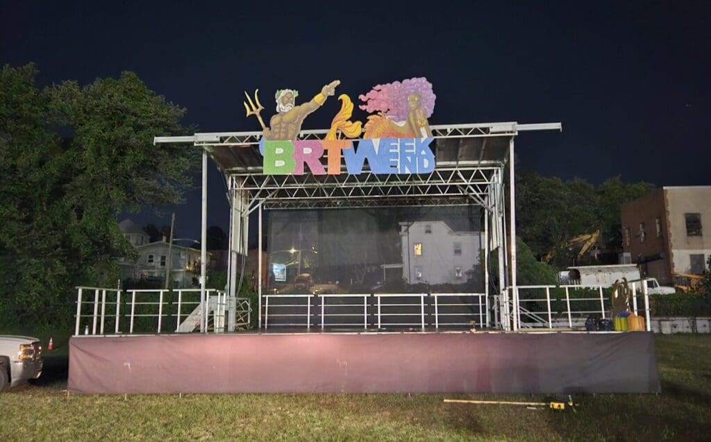 NYC portable event stage and bandshell rentals. festival in the Tri-state area provided by A view of an outdoor event with chairs and tables. An event stage with an open top and a person walking on the grass to rent in Long Island and Tri state area from Allseasonsstagerentals.com
