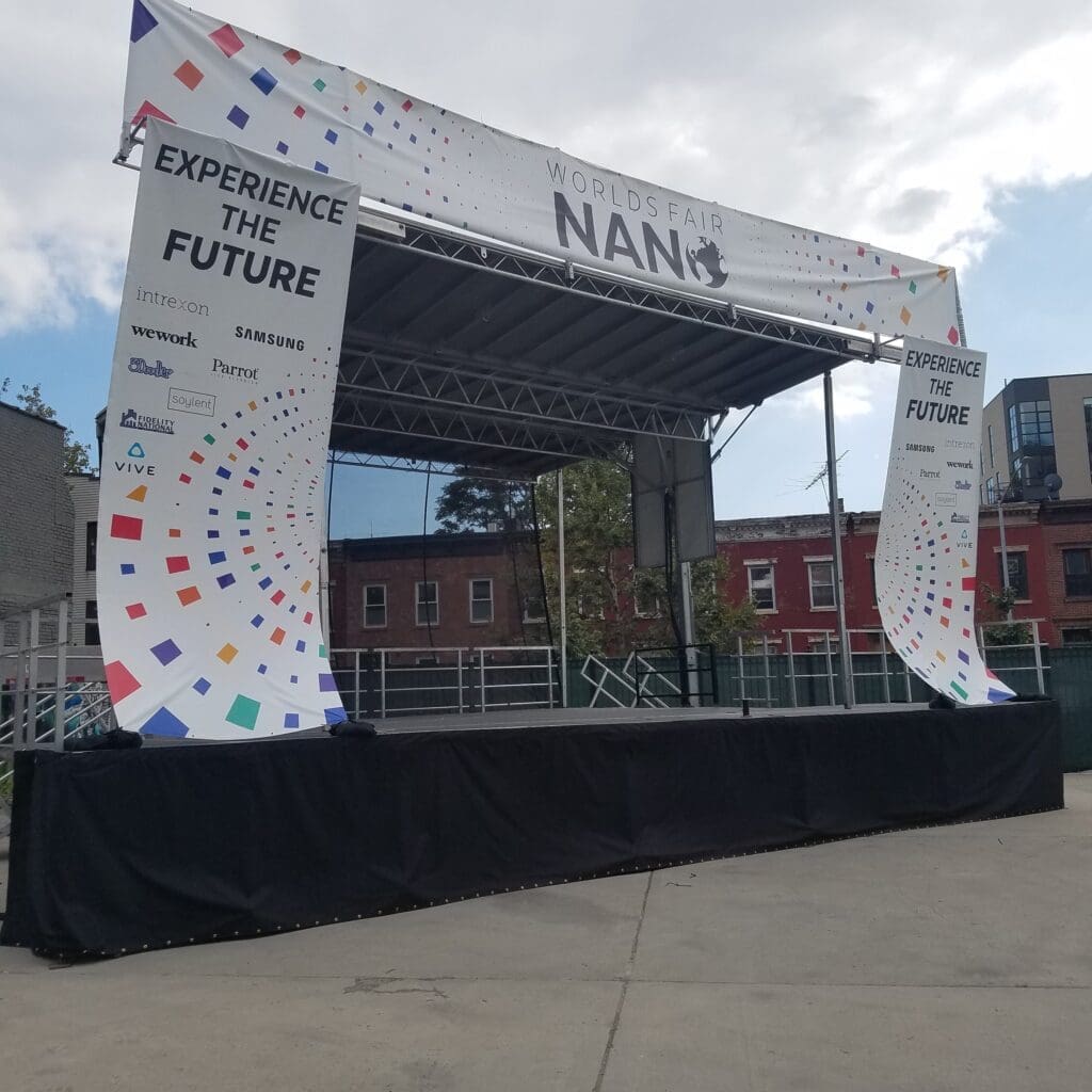 A stage with an event name on it. A view of an outdoor event with chairs and tables. An event stage with an open top and a person walking on the grass to rent in Long Island and Tri state area from Allseasonsstagerentals.com
