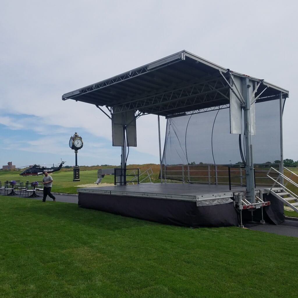 An event stage with an open top and a person walking on the grass to rent in Long Island and Tri state area from Allseasonsstagerentals.com